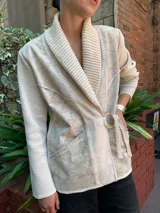 Chaqueta color crema con cuello tipo chal, cierre cruzado con cinturón y bolsillos laterales.