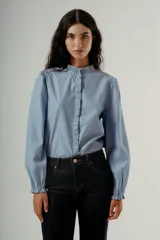 Camisa de manga larga color celeste con finas rayas verticales oscuras. Presenta cuello mao con volados y un detalle de volado vertical en el centro frontal. Las mangas tienen un ligero efecto globo en los hombros.