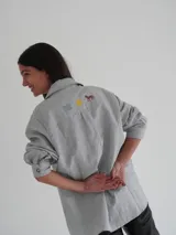 Camisa celeste a rayas verticales finas, con cuello clásico, cierre frontal con botones y mangas largas. Presenta bordados de un sol, un caballo y líneas horizontales en la parte superior de la espalda.