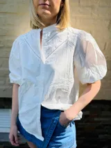 Camisa blanca de popelina de algodón, manga corta abullonada y cuello mao. Presenta doble detalle de puntilla en forma de V en la parte superior delantera.