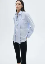Camisa celeste de popelina de algodón con lazo al cuello, cierre de botones frontal, bolsillo de parche en el pecho y mangas largas abotonadas.