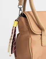 Cartera tipo city color beige, de tela deportiva, con doble asa corta, correa regulable desmontable, solapa con broche a presión y charm colgante.