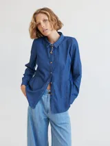 Camisa celeste de algodón con cuello clásico, cierre frontal con botones y mangas largas. Presenta volados en el frente y corte evasé.