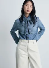 Camisa de jean celeste con cuello camisero y lazo al cuello, de manga larga con puños abotonados y corte recto.