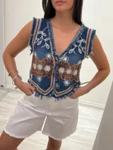 Chaleco corto de denim azul con bordados florales en blanco y marrón, detalle desflecado en escote, sisas y ruedo. Escote en V y cierre frontal con botones. Estilo boho chic.