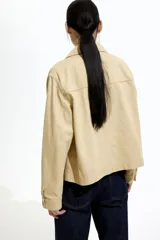 Campera color beige de sarga de algodón, con cuello, botones al frente y canesú en la espalda. Tiene hombros caídos, bolsillos superiores de parche, bolsillos insertados al frente y puños abotonados.