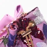 Cartera tipo saco o 'bucket bag' de tela color lila, decorada con bordados florales y lentejuelas en tonos morados, plateados y burdeos. Posee un cierre de cordón dorado con borlas de perlas y una correa de hombro de cadena dorada entrelazada con material rosa.