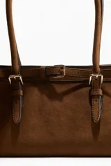Cartera marrón de gamuza sintética con doble asa corta, cierre magnético y correa decorativa con hebilla metálica.