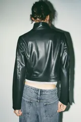 Cazadora corta negra de efecto piel, con cuello subido y manga larga. Tiene bolsillos delanteros con cremallera metálica y cierre frontal con cremallera.