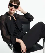 Blusa negra de chiffon con cuello alto con lazo, mangas largas con puños ajustados, detalle de volado delantero y volado en encaje.