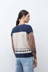 Chaqueta de tejido punto con cierre frontal, cuello redondo y manga larga. Presenta un diseño jacquard en tres colores: azul marino, beige y marrón camel. El cuerpo principal es beige con motivos de rosas azules y patrones geométricos en azul marino. Los hombros y mangas son de color marrón camel con patrones geométricos en azul marino. El cuello y el elástico inferior son de color azul marino.