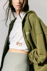 Buzo verde militar de algodón rústico con capucha, corte oversize, cierre metálico, bolsillos delanteros, estampa en frente y espalda, cordón con puntera de goma y lavado acid wash suave.
