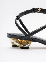 Sandalias negras de cuero con tiras finas, taco geométrico dorado y pulsera al tobillo.