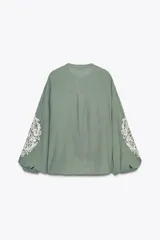 Blusa verde salvia de algodón con cuello redondo y escote pico, manga larga con encaje a contraste, puntilla y pliegues en el delantero. Cierre frontal con botones y lazada acabada en pompones.
