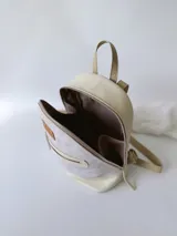 Mochila pequeña de cuero color gris claro y beige, con cierre superior y bolsillo frontal con cierre. Tiene asa de mano y correas ajustables para los hombros.