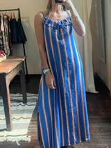 Vestido largo de tirantes finos con estampado a rayas verticales azules, blancas y marrones. Presenta escote recto con volantes y aberturas laterales.