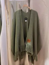 Ruana de tejido grueso color verde oliva, con cuello en V y flecos largos en el borde inferior. Presenta una etiqueta de cuero con el texto "PURA LANA" en el frente.