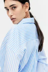 Camisa celeste y blanca a rayas verticales, de corte amplio, con cuello clásico, cierre frontal con botones, bolsillo de parche en el pecho, mangas largas con puños abotonados y bajo redondeado.