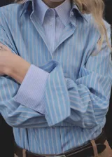 Camisa celeste a rayas verticales blancas y marrones, con cuello camisero, cierre frontal con botones, mangas largas con puños abotonados y corte recto.