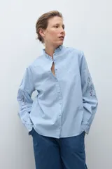 Camisa de algodón celeste claro, de manga larga, con cuello mao con volados y bordados calados florales en el frente y las mangas.