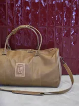 Bolso de viaje tipo duffle confeccionado en cuero genuino color camel. Posee doble asa de mano con unión de broche, correa de hombro desmontable y cierre superior. Presenta un parche frontal decorativo con el logo de la marca Enfrenada y un diseño inspirado en un sello postal uruguayo. Interior completamente forrado con bolsillo interno.