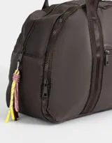 Bolso color beige de tela deportiva con manijas superiores y cierre superior. Tiene un bolsillo frontal con cierre y bolsillos laterales con cierre y tiradores de cordón y goma.