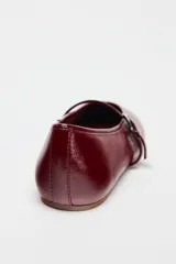 Zapato tipo bailarina color borgoña con efecto acharolado, acabado en punta redonda y tira con hebilla ajustable en la parte delantera.