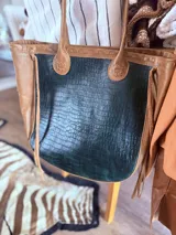 Bolso tipo tote bag con paneles laterales color cuero y panel central con textura de piel de cocodrilo color verde oscuro. Tiene doble asa de hombro color cuero y flecos laterales.