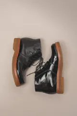 Bota de cuero vacuno con acabado charol, color marrón oscuro. Presenta un diseño de caña corta con cordones frontales y costuras decorativas en la puntera. La suela es de goma tipo crepe.