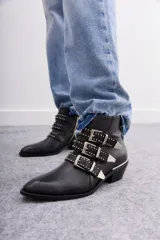 Bota de caña corta de cuero negro con tres tiras cruzadas decoradas con remaches, hebillas y puntera metálica. Tiene cierre lateral y taco ancho de 4 cm con vira metálica.