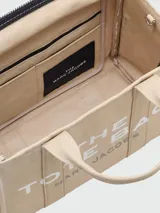 Bolso tote mediano color beige con estampado frontal del logo "The Tote Bag Marc Jacobs" en blanco. Tiene doble asa corta y correa larga extraíble de tela azul marino.