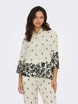 Camisa color crema con estampado de hojas y flores negras, cuello camisero, mangas 3/4 amplias y cierre frontal con botones.