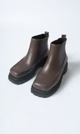 Bota corta tipo Chelsea de cuero negro, con puntera cuadrada y suela track gruesa y dentada. Presenta paneles elásticos acanalados a los lados para facilitar el calce.