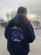 Canguro negro con capucha y cordones ajustables. Estampa en la espalda con el texto "Chema Surf Trip" y dibujos de un sol, una tabla de surf, una carpa y otros elementos relacionados con el surf.