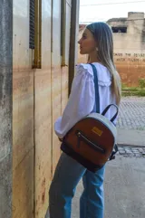 Mochila de cuero color tostado, con base y espalda en negro y tapa superior en blanco. Tiene un bolsillo frontal con cierre y un pequeño aplique de cuero con el logo de la marca.