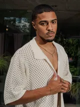 Camisa de manga corta color crudo/blanco, confeccionada en tejido de punto abierto con textura calada y cuello polo. Presenta cierre frontal de botones.