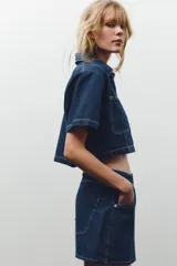 Camisa corta de denim azul con cuello solapa, escote pico, manga corta, bolsillos delanteros de plastrón y cierre frontal con botones metálicos.