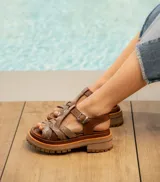 Sandalias bajas de cuero vacuno color beige con tiras cruzadas plateadas y hebilla lateral. Suela doble de goma deportiva con altura de 3cm adelante y 5,5cm atrás.