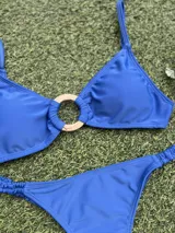 Bikini de dos piezas color azul vibrante. El corpiño es tipo top con un detalle de hebilla redonda dorada en el centro. La bombacha es colaless entera con frunces ajustables en los laterales.