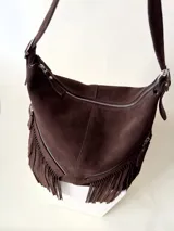 Bolso tipo hobo color tostado, hecho de cuero con flecos largos en el frente y dos bolsillos con cierre. Tiene una manija regulable con hebillas metálicas.