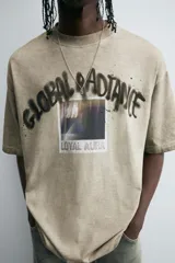 Camiseta color arena de corte holgado, manga corta y cuello redondo. Estampado frontal con la frase "Global Radiance" en estilo graffiti y una foto con efecto lavado.