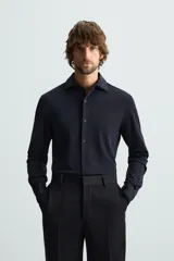 Camisa color beige de corte slim fit, confeccionada en tejido elástico que minimiza las arrugas. Tiene cuello italiano abierto, manga larga con puños abotonados y cierre frontal con botones.