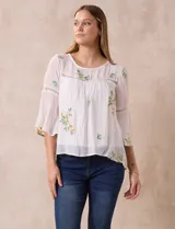 Blusa blanca de gasa con bordados florales, corte holgado, mangas 3/4 acampanadas con detalle de puntilla y forro interno.