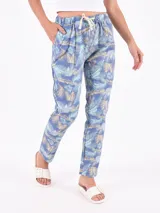Pantalón de tela estampada con hojas de palmera en tonos celeste, beige y verde sobre fondo azul. Tiene cintura elástica con cordón ajustable y bolsillos laterales.