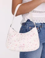 Bandolera tipo baguette de cuerina color blanco con estampado de flores pequeñas en tonos rosa y lila. Tiene manija superior, cierre superior y tag de metal.