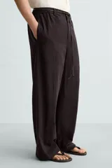 Pantalón marrón oscuro de corte ancho, confeccionado en mezcla de algodón y lino. Presenta cintura elástica, bolsillos laterales y traseros, pliegues frontales y cierre con cremallera y botón.