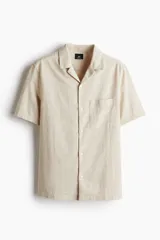 Camisa de manga corta color beige claro, confeccionada en mezcla de lino y algodón. Tiene cuello abierto, tapeta lisa, bolsillo superior abierto, canesú en la espalda y bajo recto con aberturas laterales.