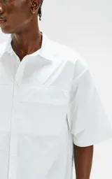 Camisa de manga corta, color blanco, confeccionada en popelín de algodón con silueta oversize. Posee dos bolsillos frontales, uno con solapa y otro con ojal y botón.