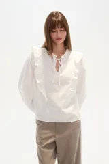 Blusa blanca de calce holgado con cuello en V y lazo frontal. Presenta detalles de volados en el pecho y cuello, y mangas largas con puños elásticos.