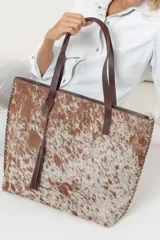 Bolso tipo tote de cuero vacuno con estampado animal print en tonos marrones y blancos. Tiene doble asa de cuero marrón, cierre superior y borla de flecos de cuero marrón.